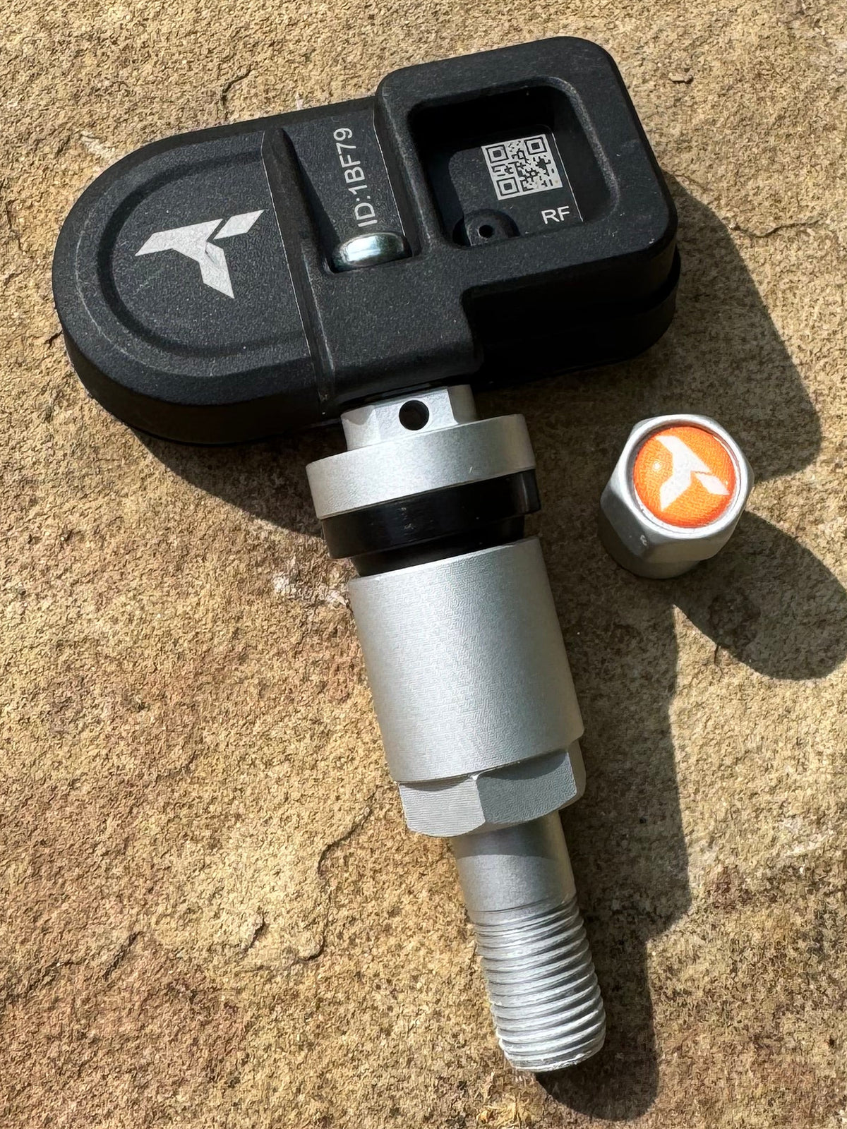 A vehicle tire pressure sensor with a black plastic housing and a silver metal threaded body, resting on a textured concrete surface.