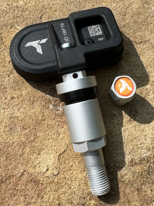 A vehicle tire pressure sensor with a black plastic housing and a silver metal threaded body, resting on a textured concrete surface.